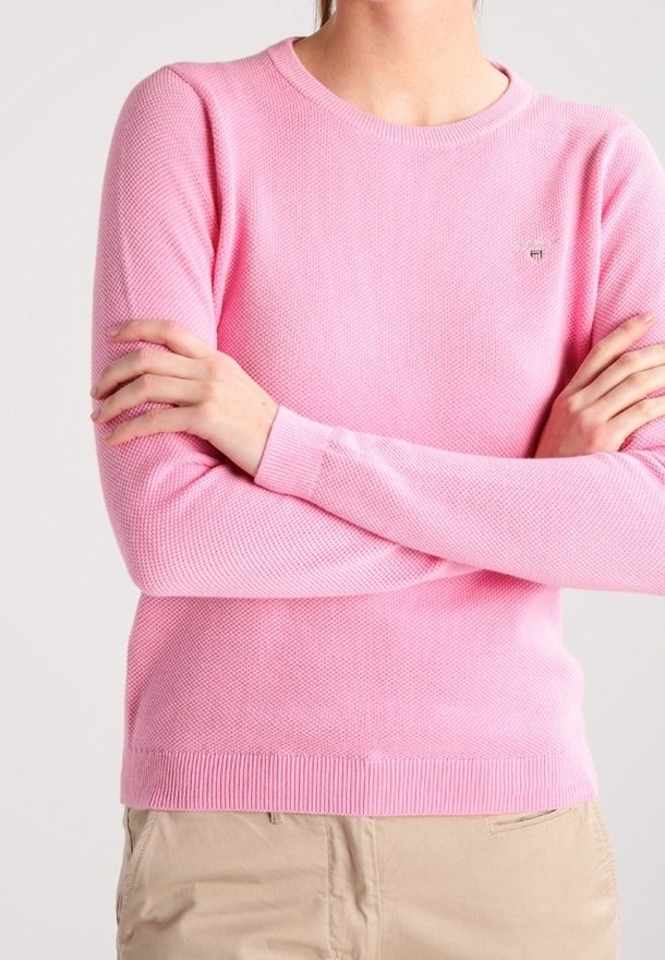 Personne portant un pull texturé rose clair à manches longues, bras croisés, associé à un pantalon beige sur un fond uni.