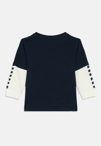 Camisa de manga longa azul-marinho com mangas inferiores brancas apresentando padrões quadrados pretos nas costas das mangas, mostrada plana por detrás.