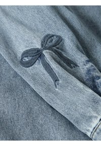 Gaiši zila džinsa auduma ar tumši zilu izšūtu loku akcentu. Tekstūra ir gluda ar izteiktām diagonālām aušanas rakstu līnijām.