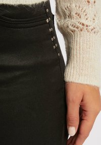 Gonna nera con una trama liscia, dettagli con bottoni laterali e un design aderente; abbinata a un leggero maglione di colore crema lavorato a maglia.
