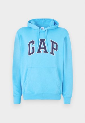 Niebieska bluza z kapturem wykonana z miękkiego materiału, z kieszenią typu kangur i sznurkiem do ściągania. Logo "GAP" w wyraźnych granatowych literach na przodzie.