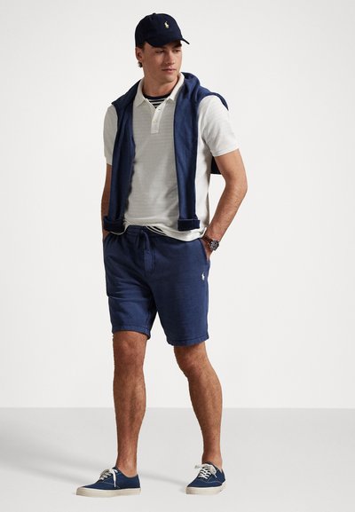 Un short bleu marine et un polo blanc, avec un sweats bleu marine drapé sur les épaules et une casquette assortie. Des baskets en toile complètent le look.