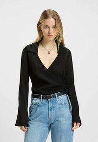 Sort indpakningsbluse med dyb V-udskæring, lange flagrende ærmer og tekstureret stof; parret med lyseblå højtaljede jeans og et sort bælte.