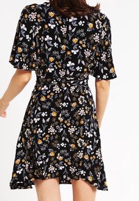 Mujer vistiendo un vestido negro con flores amarillas, blancas y lavanda, de mangas cortas y cintura ajustada, vista desde atrás.