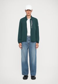 Σκούρο πράσινο μπουφάν με φερμουάρ και τσέπη στο στήθος, φορεμένο πάνω από λευκό μπλουζάκι και χαλαρά ανοιχτό μπλε τζιν. Μαύρα παπούτσια ολοκληρώνουν το ντύσιμο.