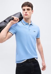 Νεαρός άνδρας με ανοιχτόχρωμο μπλε πόλο πουκάμισο κρατάει μια μαύρη δερμάτινη τσάντα στον ώμο του, με απλό ανοιχτόχρωμο φόντο.