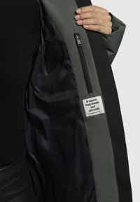 Zwarte en groene jas met een gladde binnenkant, voorzien van een ritsvak en een label met de tekst "Een warme omhelzing gemaakt van stof."