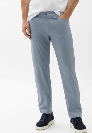 Mand iført lyseblå teksturerede bukser, hvid t-shirt og marineblå sneakers med hvide såler, stående med hænderne i lommerne.