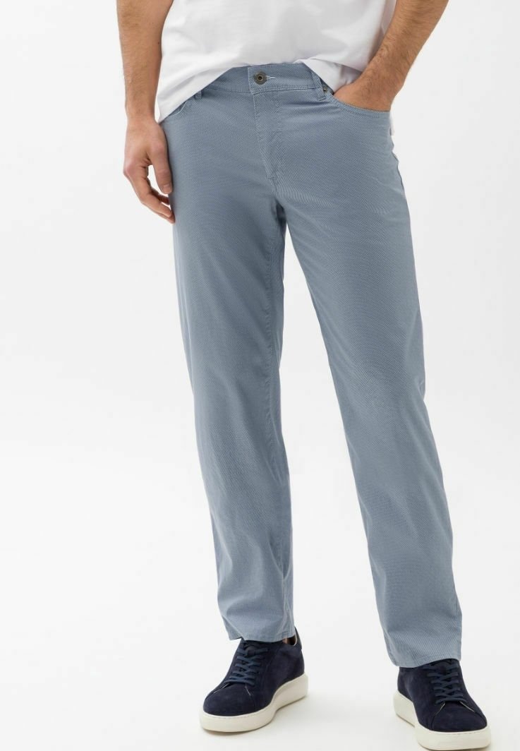 Homme portant un pantalon bleu clair texturé, un t-shirt blanc et des baskets bleu marine avec des semelles blanches, debout les mains dans les poches.