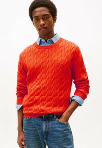 Maglione arancione a maglia rasata con polsini e scollo a coste, indossato sopra una camicia azzurra con bottoni, abbinato a jeans blu.