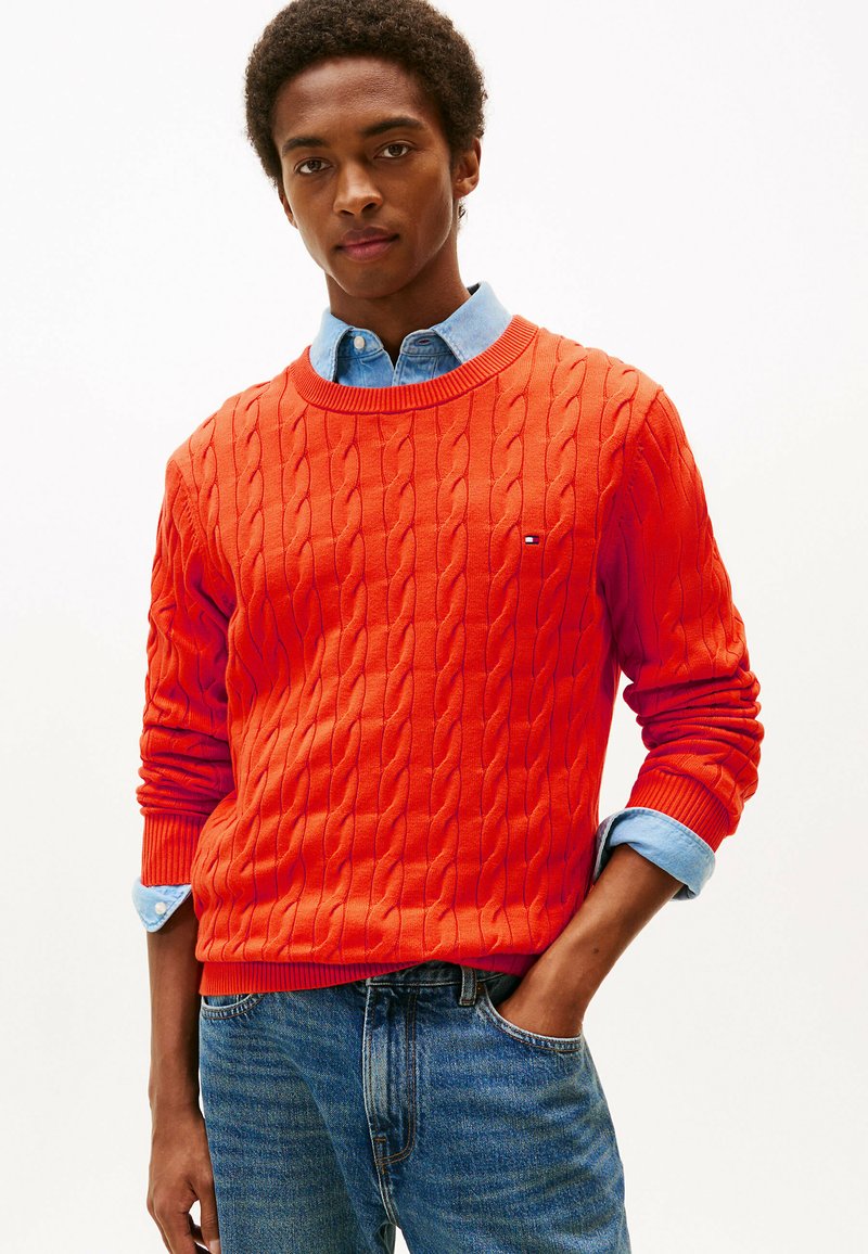 Maglione arancione a maglia rasata con polsini e scollo a coste, indossato sopra una camicia azzurra con bottoni, abbinato a jeans blu.