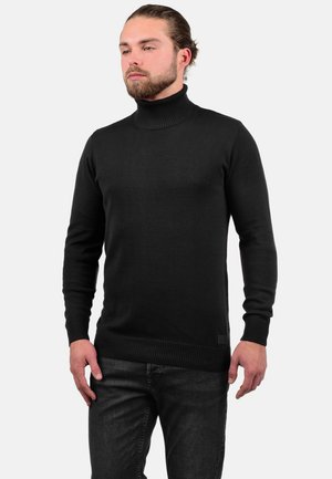 Suéter negro de cuello alto en textura de punto acanalado, con mangas largas y un diseño ajustado, combinado con pantalones vaqueros oscuros.