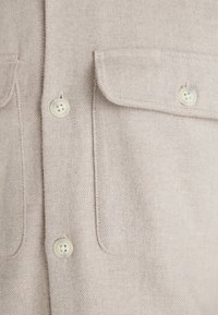 Jasnobeżowa koszula zapinana na guziki wykonana z teksturowanego materiału. Posiada dwie kieszenie na piersi, zaokrąglony kołnierz oraz cztery jasne guziki.