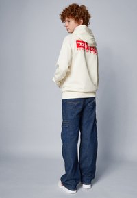 Felpa beige con logo rosso sul retro, abbinata a jeans cargo blu scuro con tasche. Il modello è in piedi di lato su uno sfondo neutro.