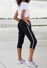 Pantalon de sport noir court avec bandes blanches sur les côtés, taille élastique et poches, associé à un t-shirt blanc noué et des baskets grises.