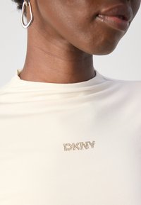 Nærbilde av en person som har på seg en kremfarget topp med små gull DKNY-bokstaver nær halsutskjæringen og en sølvdråpeøredobbe.