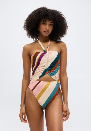 Kobieta o kręconych włosach w wielobarwnym, paskowanym kostiumie jednoczęściowym z marszczeniami i wycięciami, stojąca na jednolitym tle.