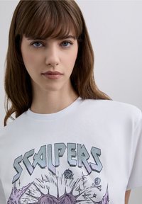 Wit katoenen t-shirt met een grafische print met paarse en grijze tekst "SCALPERS" en illustraties van uitbarstingen en schedels.