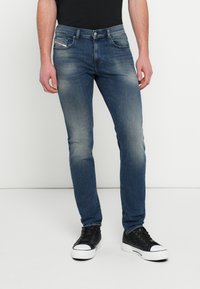 Skinny Fit Denim-Jeans in mittlerem Blau, mit leichtem Used-Effekt, klassischem Fünf-Taschen-Design und Metallknopfverschluss. Kombiniert mit schwarzen Sneakers.