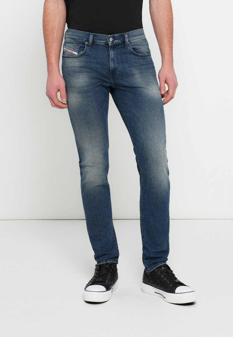 Skinny Fit Denim-Jeans in mittlerem Blau, mit leichtem Used-Effekt, klassischem Fünf-Taschen-Design und Metallknopfverschluss. Kombiniert mit schwarzen Sneakers.