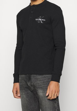 Mann trägt ein schwarzes Calvin Klein Jeans Langarmshirt mit Knöpfen am Kragen und ausgewaschene schwarze Jeans.
