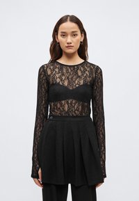 Μαύρο μπλουζάκι με μακριά μανίκια από δαντέλα με διαφανές σχέδιο και μαύρο σουτιέν από κάτω. Συνδυασμένο με μαύρη πλισέ φούστα με λεπτομέρεια κουμπιών.