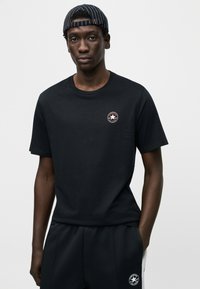 Μαύρο βαμβακερό t-shirt με κοντά μανίκια και στρογγυλή λαιμόκοψη. Διαθέτει έν α μικρό στρογγυλό λογότυπο στο στήθος. Το ύφασμα φαίνεται λείο και αεριζόμενο.