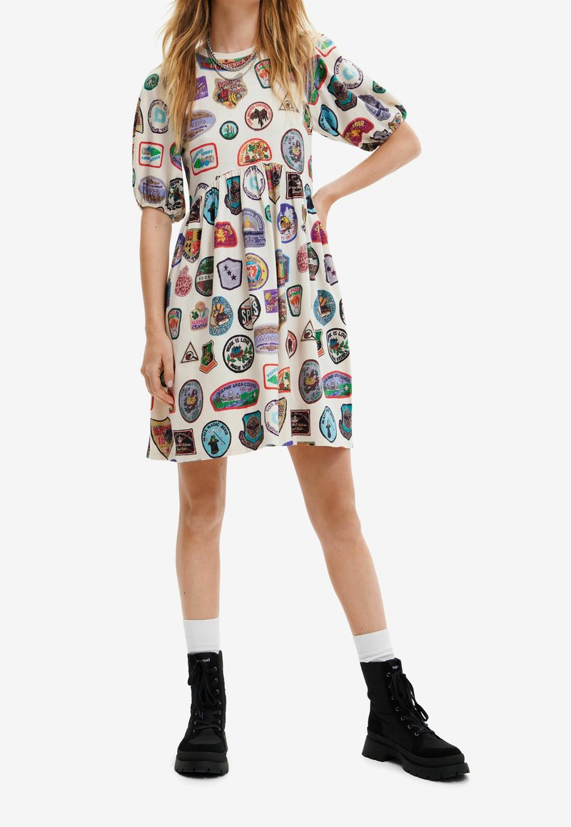 Femme portant une robe blanche à manches courtes avec des patchs colorés, associée à des chaussettes blanches et des bottines noires à lacets.