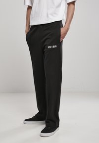 Sorte sweatpants med en afslappet pasform, der har hvid broderet tekst på venstre lår og sidelommer, parret med mørke sneakers.