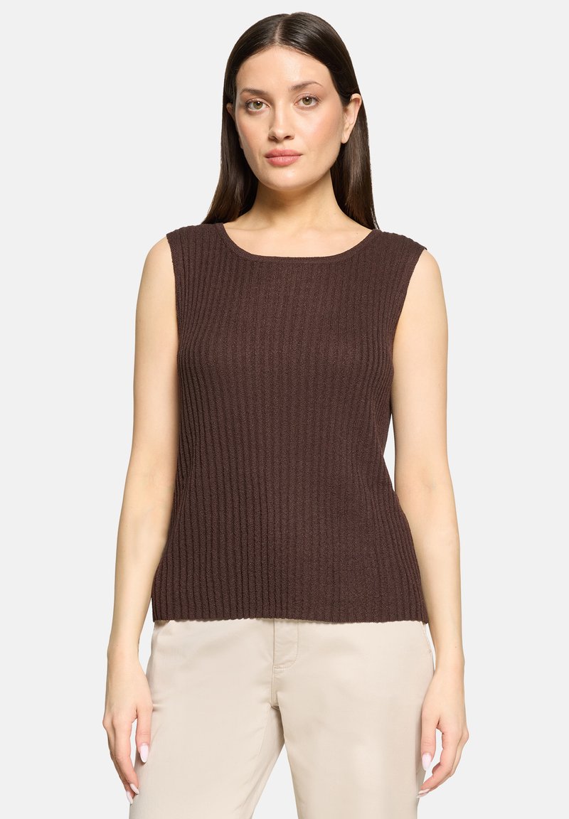 Femme aux cheveux raides et foncés portant un haut marron sans manches côtelé et un pantalon beige, debout devant un fond clair uni.