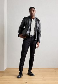 Veste en cuir noire sur un henley gris, assortie à un pantalon et des chaussures noirs. Tient un sac à bandoulière noir. Design simple et épuré sans motifs.