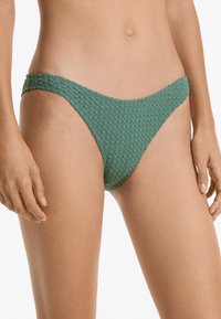 Zelené bikiny s nízkým pasem, vyrobené z texturované tkaniny s mřížkovým vzorem, které mají méně krytí na bocích. Měkký, pletený materiál.