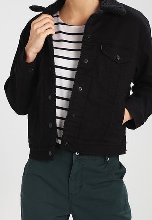 Person wearing a black buttoned jacket with fleece collar over a white and black striped shirt and dark green pants, adjusting jacket collar.