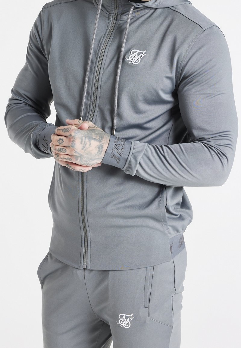 Haut de sweatshirt zippé gris en tissu lisse avec un logo sur la poitrine. Présente un design ajusté et un cordon de serrage ajustable à la capuche.