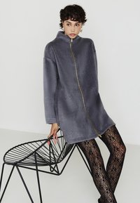 Una persona giovane con capelli ricci e corti indossa un ampio cappotto grigio con zip, collant neri in pizzo floreale e orecchini a cerchio dorati, mentre si appoggia a una sedia di metallo nera.