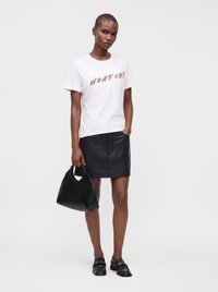 Γυναίκα που φοράει λευκό γραφικό t-shirt, μαύρη δερμάτινη φούστα με κουμπιά, μαύρα loafers και κρατάει μαύρη τσάντα, στέκεται με το ένα χέρι στην τσέπη.