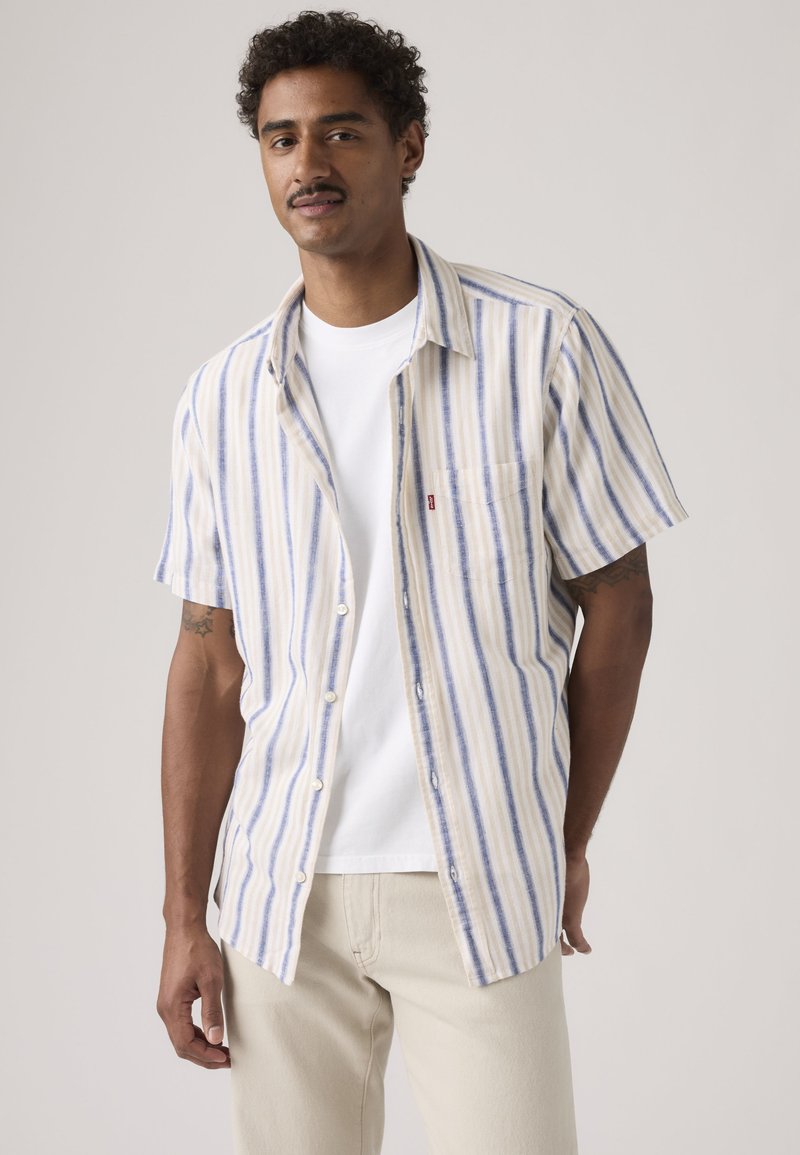 Mand med kort krøllet hår iført en beige og blåstribet kortærmet skjorte over en hvid t-shirt og lyse beige bukser, stående mod en ensfarvet baggrund.