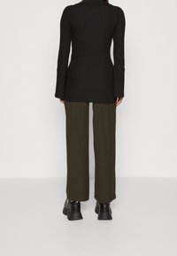 Person som står med ryggen mot kameran, iklädd en ribbad svart långärmad topp, olivgröna vida byxor och svarta klackskor med tjock sula på en grå golv.