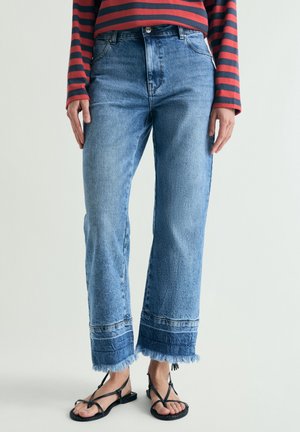 Personne portant un jean bleu taille haute avec des ourlets effilochés et des sandales noires à lanières, assorti à un haut à manches longues rayé rouge et bleu marine.