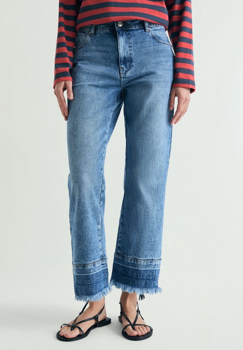 Personne portant un jean bleu taille haute avec des ourlets effilochés et des sandales noires à lanières, assorti à un haut à manches longues rayé rouge et bleu marine.