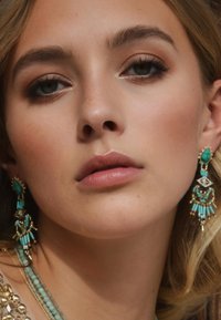 Boucles d'oreilles en perles teal avec des accents en or présentent une forme en goutte d'eau, des détails complexes et des éléments de franges superposées, mettant en valeur des couleurs vives.