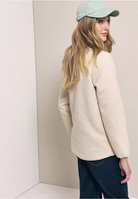 Veste en polaire beige clair avec une surface texturée, col rond et manches longues. Portée avec une casquette vert menthe, vue de dos.