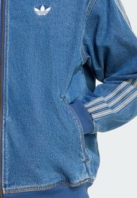 Jeansjacke in Hellblau mit einem Reißverschluss vorne, gerippten Bündchen und drei weißen Streifen an den Ärmeln. Kleines weißes Logo auf der Brust.