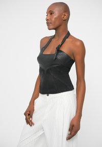 Mulher vestindo um top halter preto com detalhe de fivela e calças brancas de risca de giz, posando de lado contra um fundo simples.