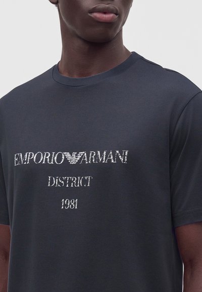 Άνδρας που φοράει σκούρο γκρι T-shirt με λευκό κείμενο ""EMPORIO ARMANI DISTRICT 1981"" στο μπροστινό μέρος.