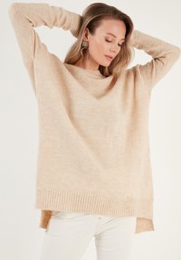 Pull en tricot beige avec une coupe ample et oversize, ourlet côtelé et fentes latérales. Texture douce et encolure large pour plus de confort.