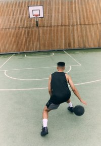 Basketballspiller dribler en sort bold på en grøn bane. En trævæg og en kurv er synlige i baggrunden. Han har en sort tanktop og grå shorts på.