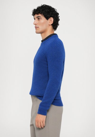 Modra pletena sv sweater s okroglo ovratnikom in dolgimi rokavi, ki ima teksturirano površino in oprijet kroj. Nosimo ga z svetlo sivimi hlačami.