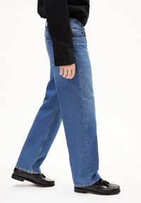 Jean en denim bleu avec une coupe droite, présentant un design classique à cinq poches et des accents cousus, associés à des chaussures en cuir noires.