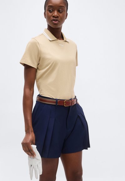 Γυναίκα που φοράει μπεζ πόλο μπλούζα και σκούρο μπλε πλισέ σορτς με καφέ ζώνη, κρατώντας ένα λευκό γάντι του γκολφ στο δεξί της χέρι.